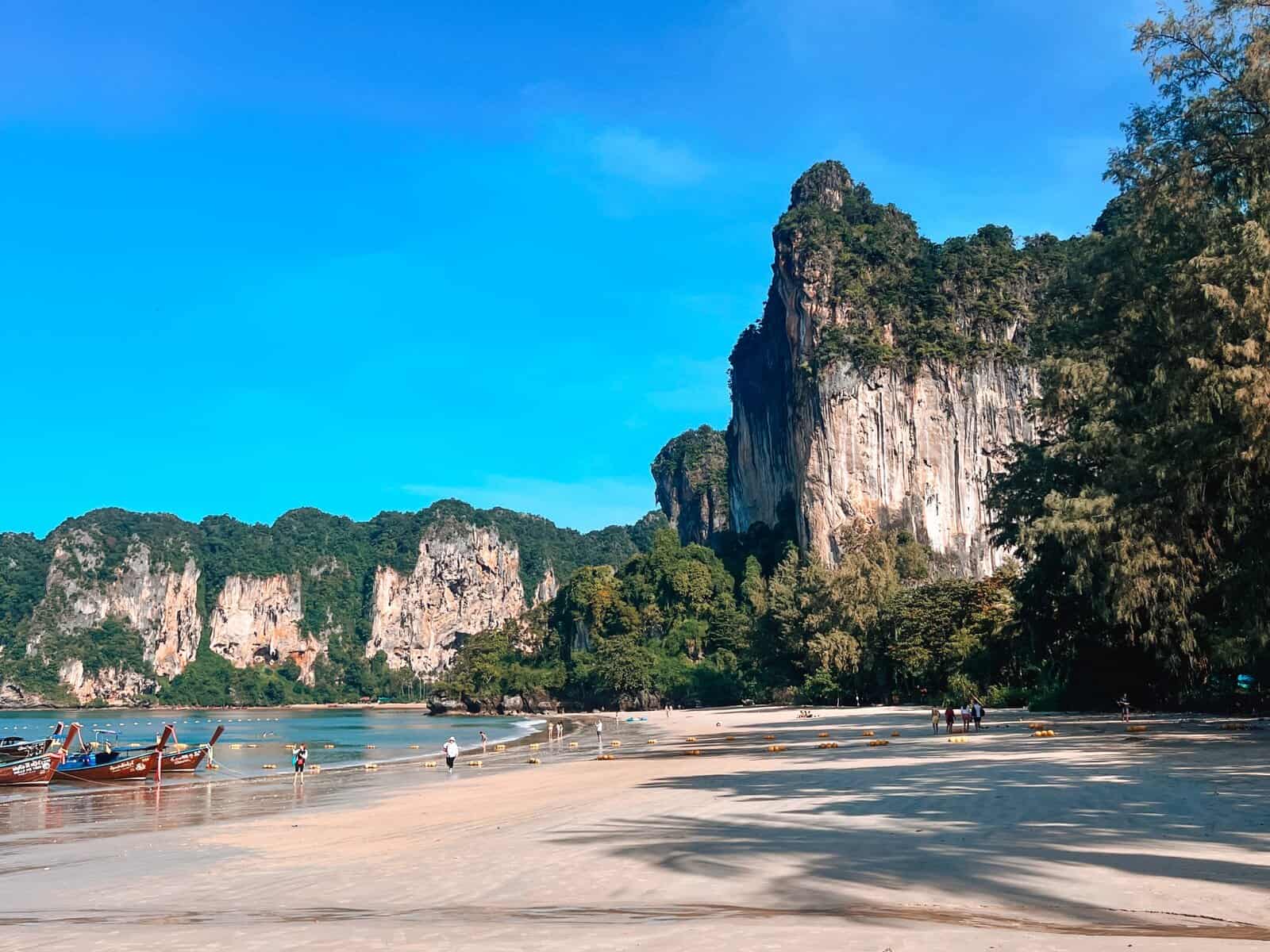 Turquoise water and limestone cliffs rise above Railay Beach in southern Thailand with longtail boats anchored along the shore and visitors walking the wide sandy beach. Although Railay Beach is well known it is featured in this hidden gems in Thailand guide because it is only accessible by boat making the journey feel secluded and memorable despite its popularity.