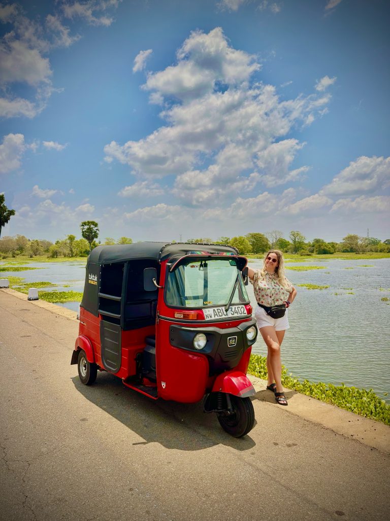Blonde traveler standing beside a red tuk tuk by a lake in Sri Lanka under the midday sun.