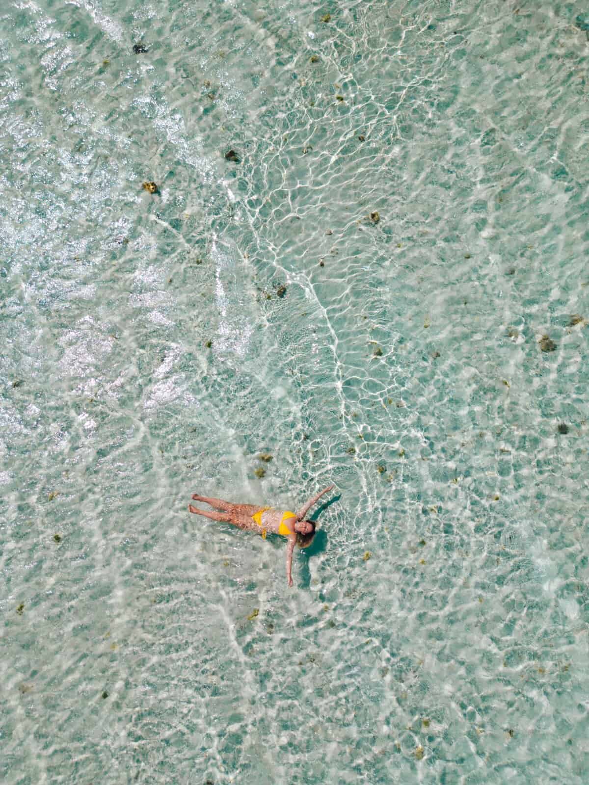 Drone shot of a woman floating on her back in clear shallow turquoise water over the White Island sandbank in Camiguin. The rippled seabed and scattered sand patches show the calm open seascape that makes White Island a popular stop in a Camiguin travel guide. This image highlights the relaxed island experience often featured in Camiguin travel and tourist places content.