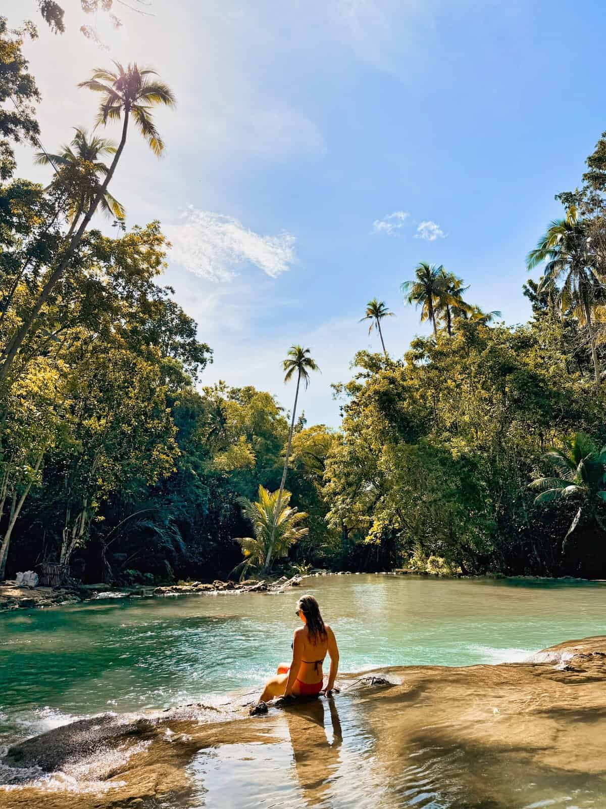 A woman sits on smooth rock at the edge of a clear turquoise waterfall pool surrounded by dense tropical forest and tall palm trees. Sunlight filters through the canopy creating a peaceful jungle swimming spot that shows the natural beauty of Siquijor.