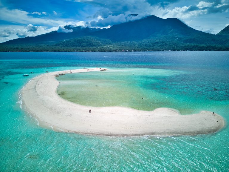 Aerial view of White Island in Camiguin showing a curved white sandbank surrounded by shallow turquoise water with Camiguin Island and its green mountains in the background. Small boats and visitors are scattered along the sandbar highlighting this popular Camiguin travel attraction accessible by boat.