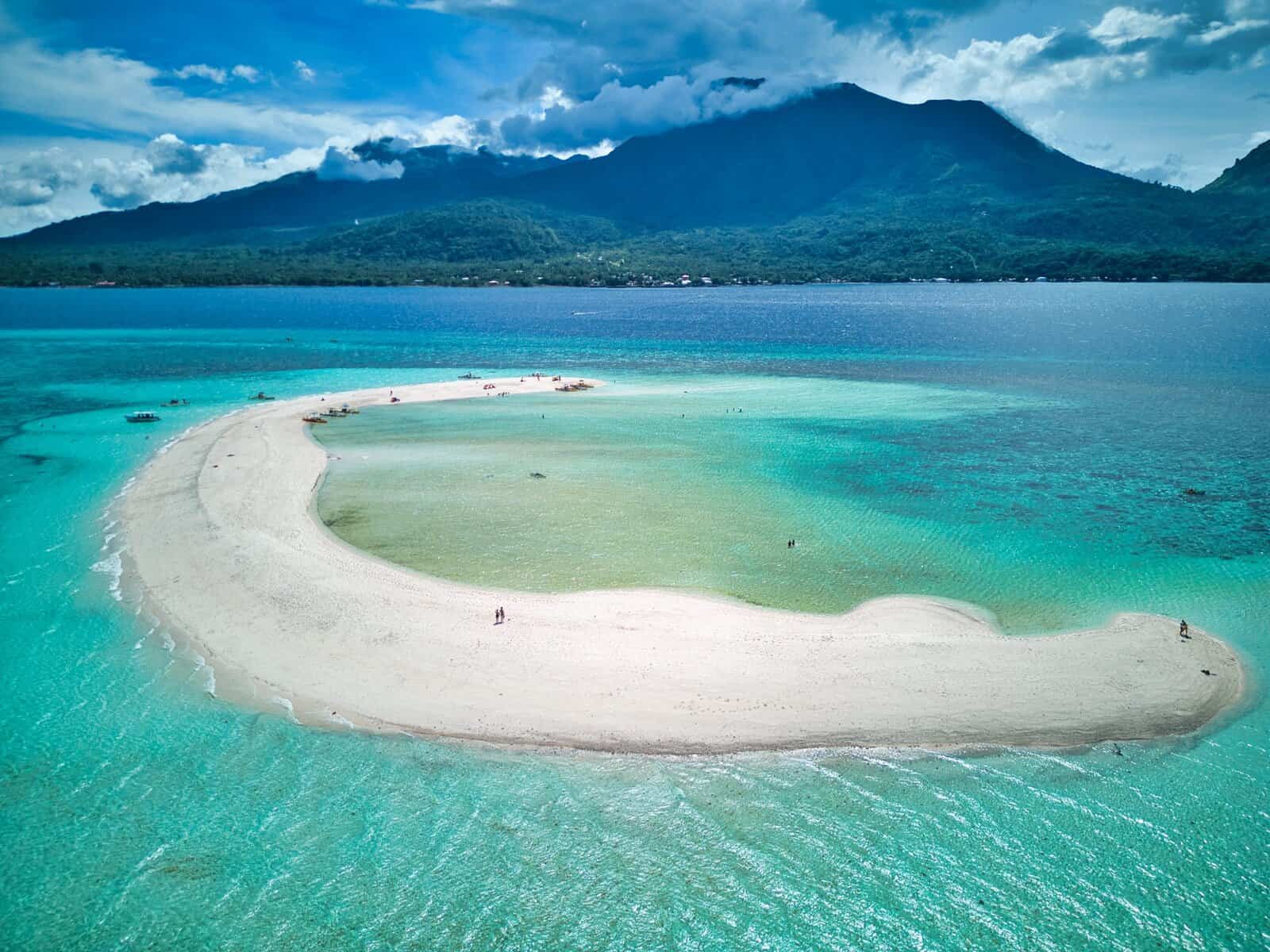 Aerial view of White Island in Camiguin showing a curved white sandbank surrounded by shallow turquoise water with Camiguin Island and its green mountains in the background. Small boats and visitors are scattered along the sandbar highlighting this popular Camiguin travel attraction accessible by boat.