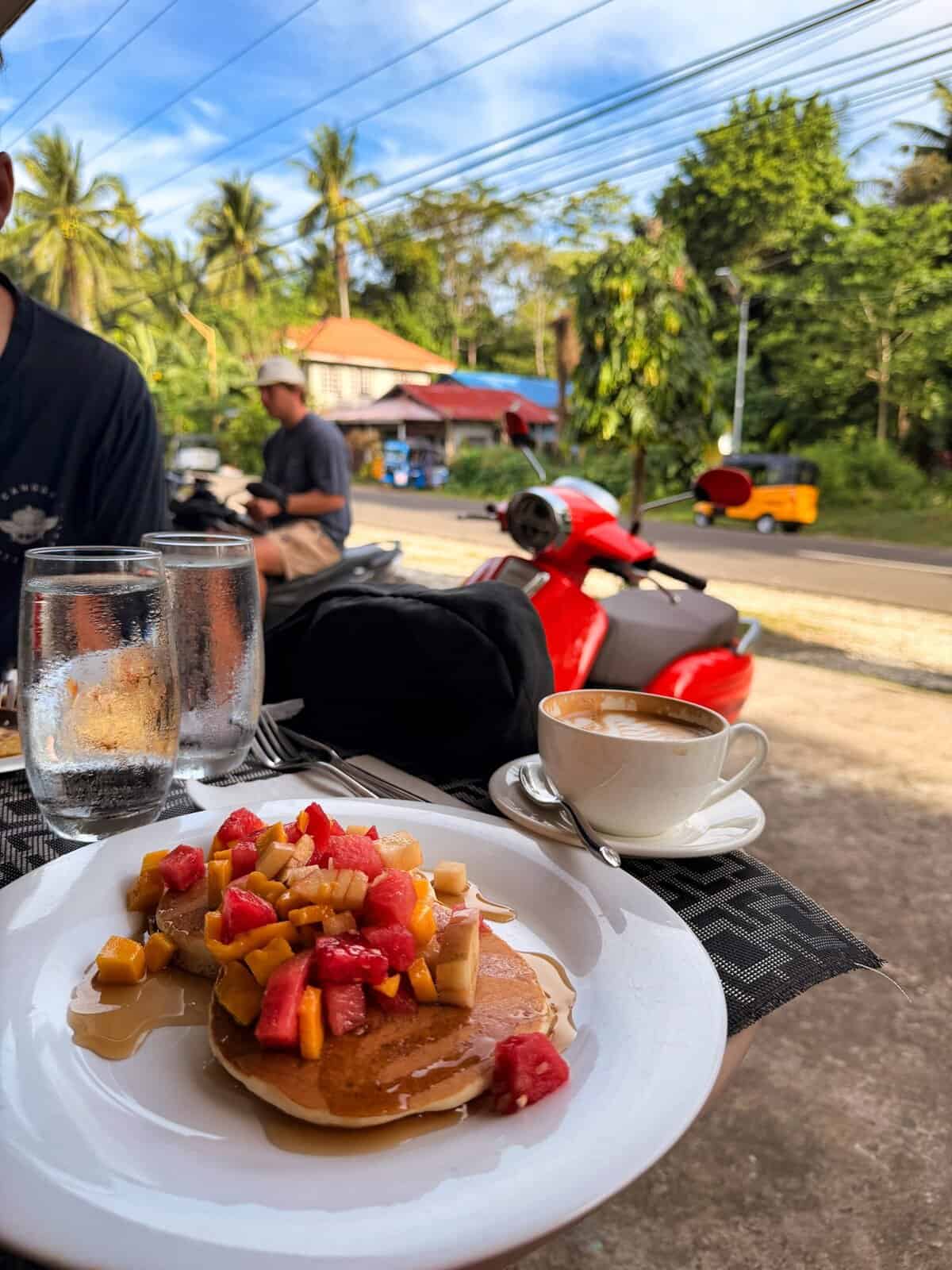 Pancakes topped with fresh tropical fruit and syrup sit on a table at Fig Cafe in San Juan, Siquijor, with coffee and water beside the plate. A red scooter and quiet roadside scene in the background capture the relaxed island vibe of a Siquijor morning.