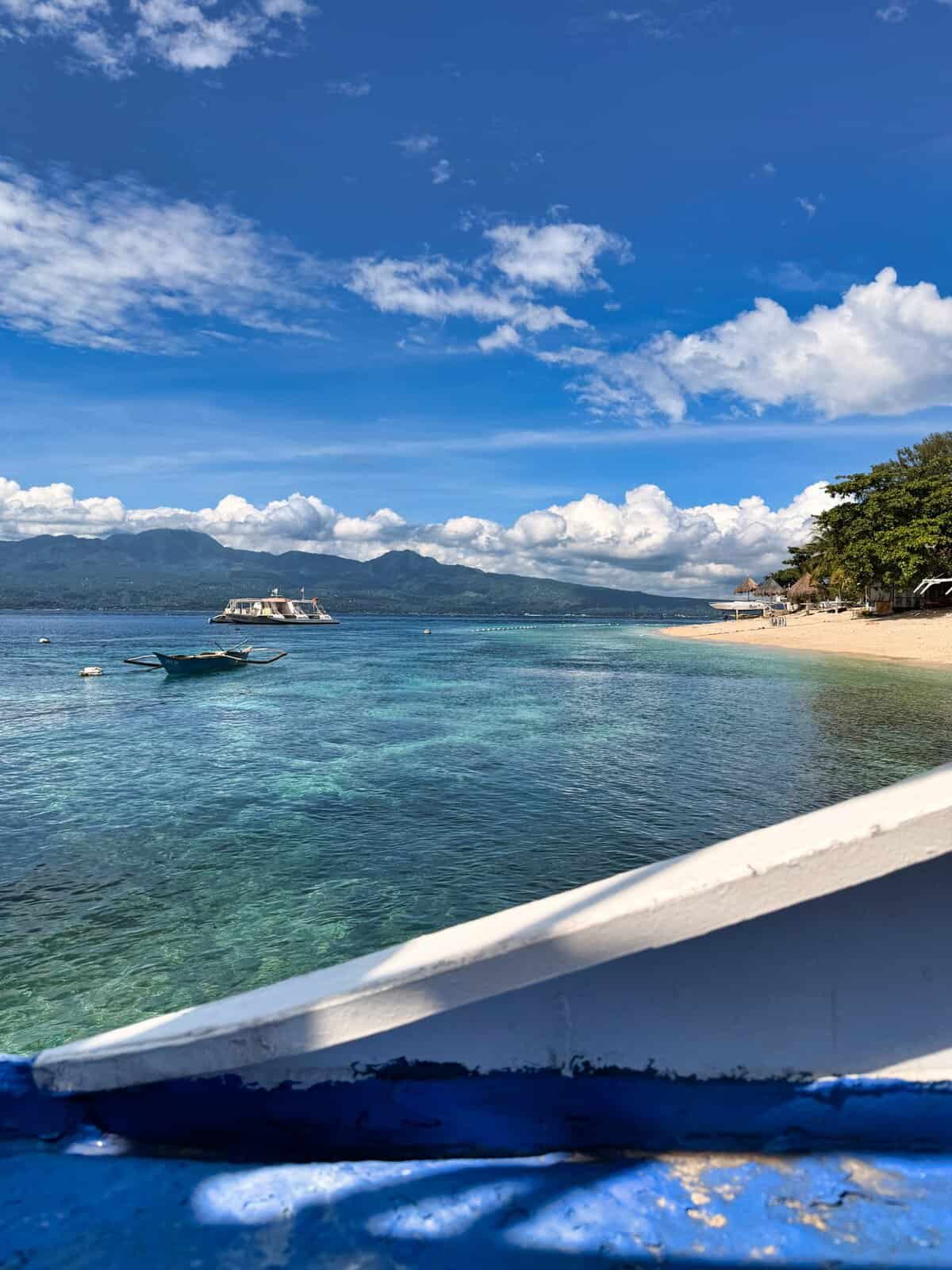 View from a boat at Liloan Port in Cebu showing calm blue water a small outrigger boat and a larger ferry with mountains in the distance. This image illustrates what travelers can expect when taking the ferry route from Cebu to Siquijor.