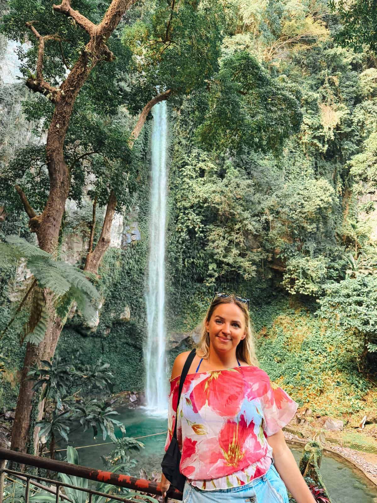 Katibawasan Falls cascades in a tall narrow stream into a clear blue pool framed by lush green cliffs. A woman stands at the viewpoint showing why Camiguin tourist places are nature spots like this waterfall.