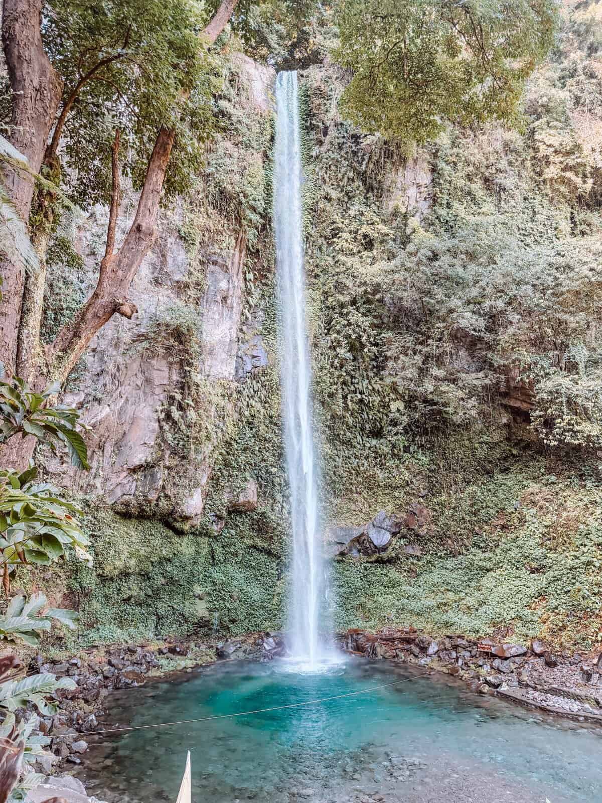 Katibawasan Falls cascades in a tall narrow stream into a clear blue pool surrounded by lush green cliffs. This waterfall highlights a must see natural attraction featured in any Camiguin travel guide.