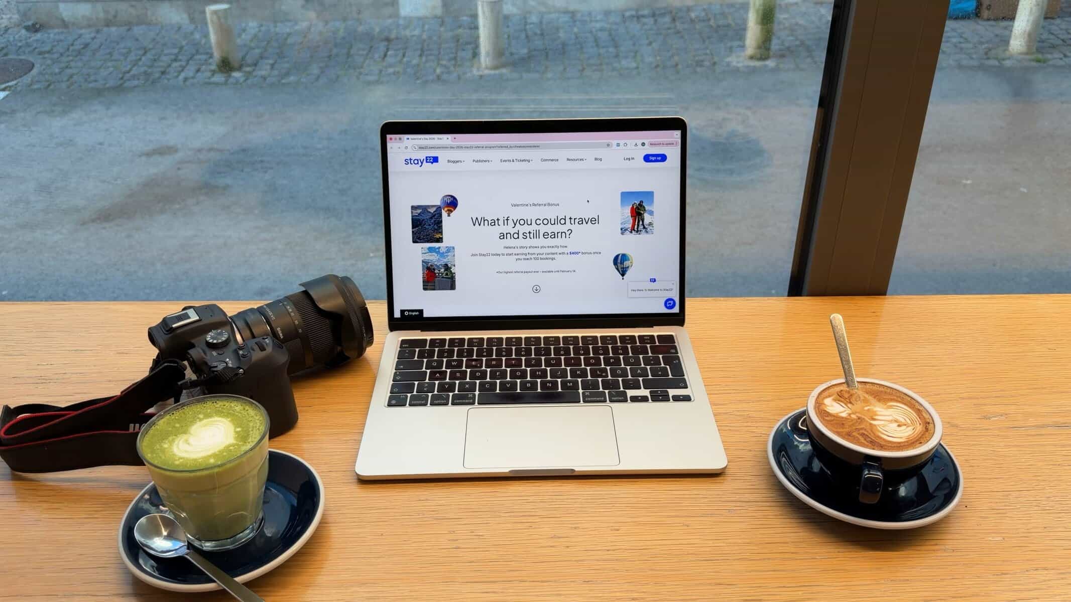 Stay22 affiliate platform homepage displayed on MacBook laptop showing "What if you could travel and still earn" with travel blog monetization value proposition – Remote work from cafe with coffee