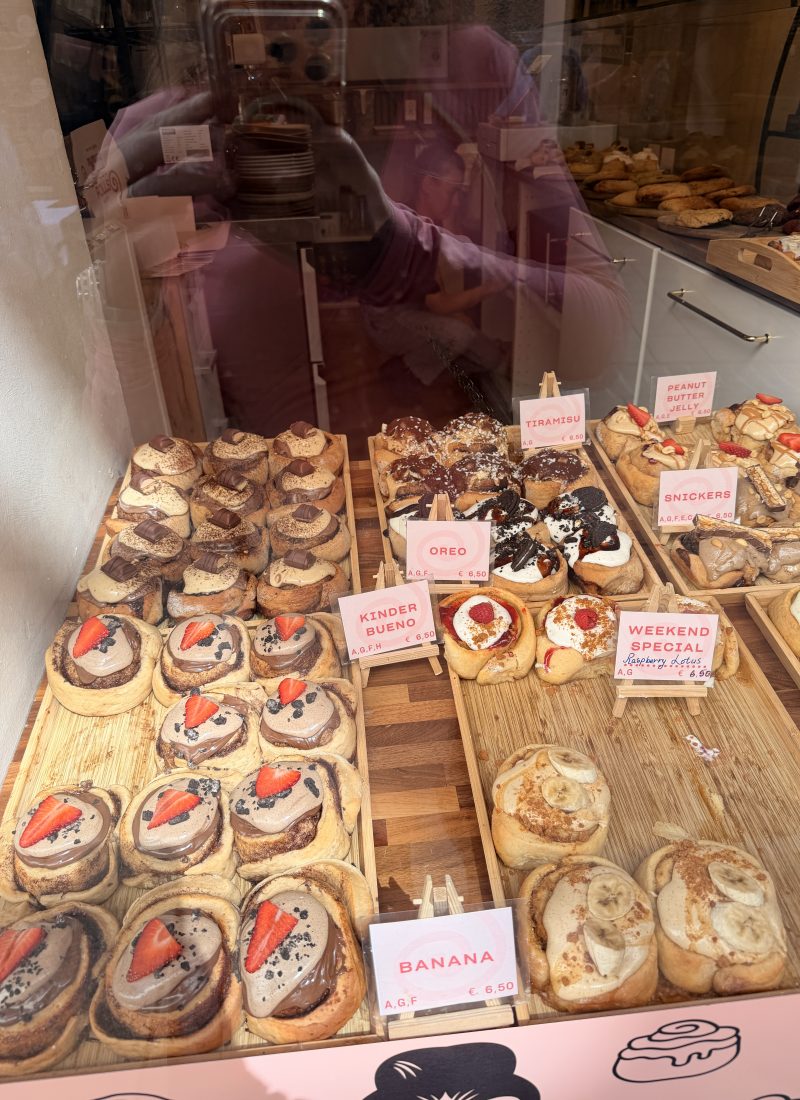 Assorted gourmet cinnamon rolls topped with Oreo, Kinder Bueno, Snickers, banana, and fresh strawberries in a bakery window in Salzburg.