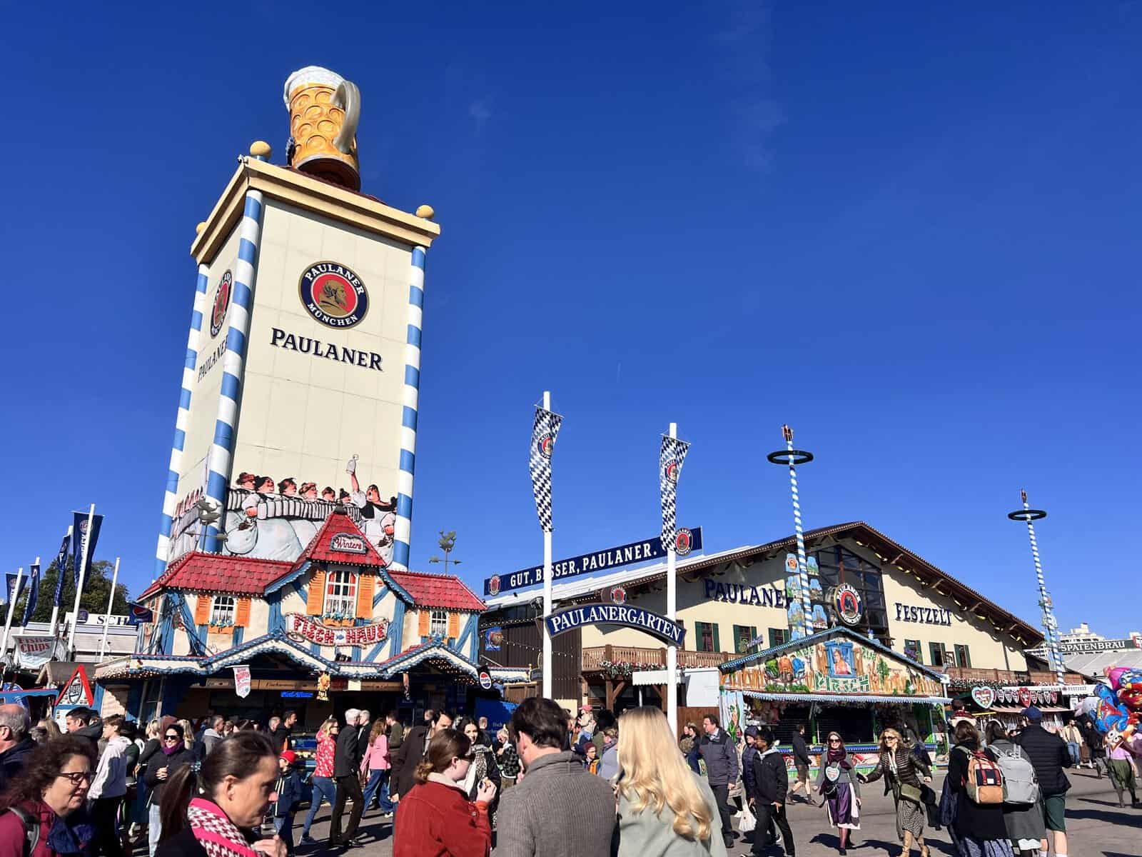 Crowds outside the Paulaner Festzelt beer tent at Oktoberfest in Munich, beneath its giant beer mug tower and a clear blue sky.