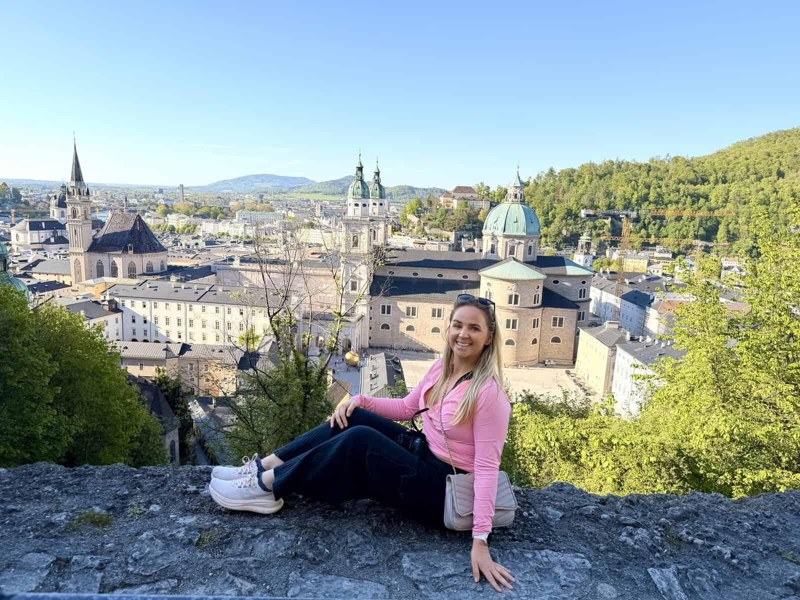 A woman sits at a rocky viewpoint above Salzburg’s Old Town, overlooking Salzburg Cathedral, DomQuartier, church towers, and green hills on a sunny day.
